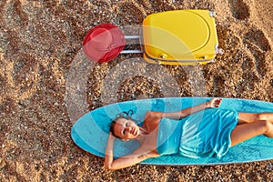 Desire of adventure, summer time, surf and holiday trip idea. Cute woman lay on surfboard, suitcase and hat on sand