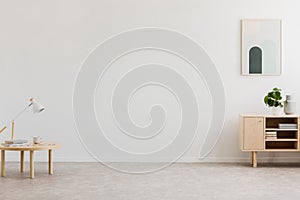 Desk lamp on a small table and a simple, wooden cabinet in an empty living room interior with white wall and place for a sofa. Rea