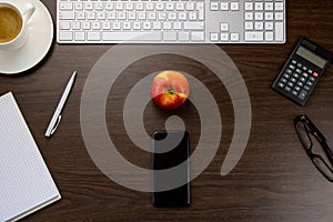Desk in the office with a smartphone and a red apple in the middle, a notepad and pen, a keyboard next to a cup of coffee and a c