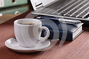 Desktop with coffee cup, opened laptop computer, diary, pan and document folders on background, no people, focused on coffee