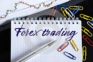 On the desktop are a forex chart, paper clips, a pen and a notebook in which it is written - Forex trading