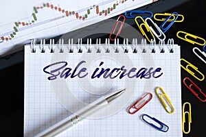 On the desktop are a forex chart, paper clips, a pen and a notebook in which it is written - Sale increase