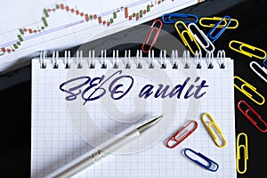 On the desktop are a forex chart, paper clips, a pen and a notebook in which it is written - SEO audit