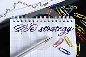 On the desktop are a forex chart, paper clips, a pen and a notebook in which it is written - SEO strategy
