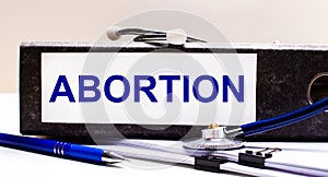 The desktop has a stethoscope, a blue pen, and a gray file folder with the text ABORTION. Medical concept