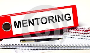 On the desktop are notebooks, a pen and a red folder for papers with the text MENTORING. Business concept