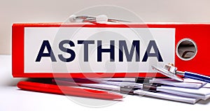 On the desktop is a stethoscope, documents, a pen, and a red file folder with the text ASTHMA. Medical concept