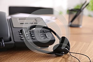 Desktop telephone and headset on wooden table in office, closeup. Hotline service