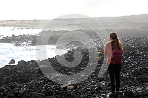 Desolate volcanic black rocks with lonely girl. Back view of young woman standing reflexive and sad on hidden rocky beach. Young