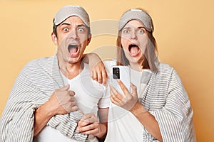 Despair shocked couple man and woman wrapped in blanket isolated over beige background looking at clock on phone being late