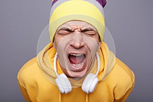 Despair stressed hipster man wearing yellow hoodie and beanie hat, keeps eyes closed, screaming loud, being depressed, has serious