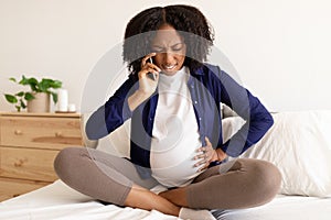 Despaired curly pregnant millennial black woman in domestic clothes with big belly calls by phone