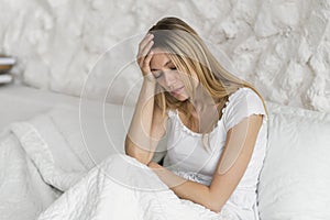 Despaired millennial caucasian blonde woman sit on bed presses hand to head, suffers from migraine