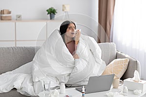 Despaired sad young indian lady wrapped in blanket sneezes in napkin, studies and works at home in living room interior