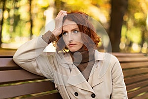 Despaired unhappy young european red-haired lady in raincoat suffers from headache, loneliness sits on bench