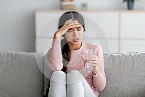 Despaired upset young indian lady suffer from fever and headache on sofa with glass of water in living room