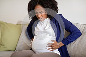 Despaired young african american pregnant woman sit on sofa suffering from contractions, screaming