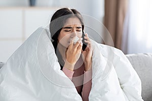 Despaired young indian female wrapped in blanket blows nose in napkin, sneeze, calls by phone on couch
