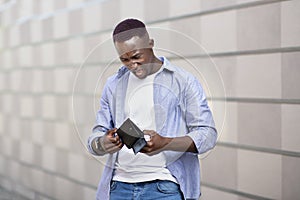 Desperate African American guy looking at his empty wallet near brick wall on city street