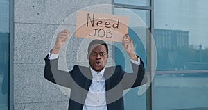Desperate african american man in formal suit holding placard NEED JOB and shouting near office building, tracking shot
