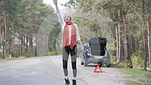 Desperate blond woman hitchhiking on suburban road. Wide shot portrait of stressed Caucasian female driver having car