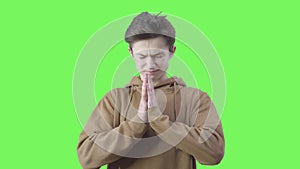 Desperate boy praying at green background. Portrait of teenage brunette Caucasian believer with hands clasped for