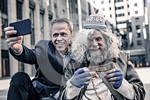 Desperate poor old man kissing dollar bill while smiling man making photo