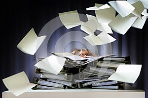 Desperate woman sinks behind stacks of ring binders on an office desk and lots of papers are flying down, concept of excessive