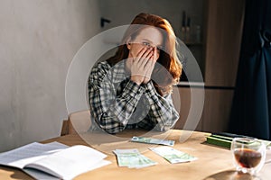 Desperate young woman counting money with sad expression, sitting at home kitchen table. Sad female having financial