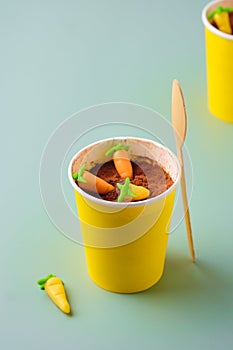 Dessert, carrot cake to go in a yellow paper cup, decorated with cocoa and marzipan carrots on a green plain background. Trendy