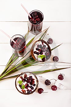 Dessert of cream and berries of currants and cherries in coconut cups, cherry juice with ice in glasses stand on a wooden white