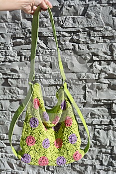 Detail of beautiful colorful crocheted handbag with green handle in the left hand behind the gray stone wall in the garden