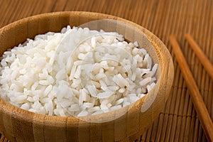 Detail of cooked white rice in a dark wood bowl next to chopsticks on bamboo matt
