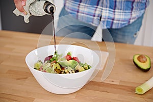 Detail on the hands of chef pouring balsamic vinegar into a bowl of fresh salad while cooking in minimalist home kitchen
