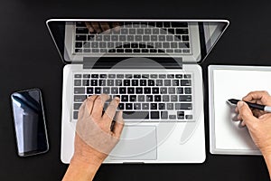 Detail of hands working on a laptop and a graphic tablet with a cell phone on black background