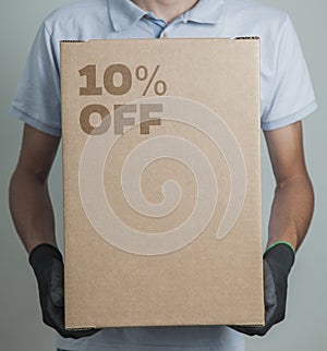 Detail of man with black gloves holding a cardboard box with 10% discount for delivering products on gray background.