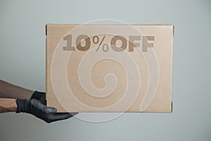 Detail of man with black gloves holding a cardboard box with 10% discount for delivering products on gray background.