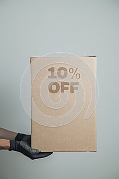 Detail of man with black gloves holding a cardboard box with 10% discount for delivering products on gray background.