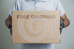 Detail of man with black gloves holding a cardboard box written free shipping to deliver products on gray background.