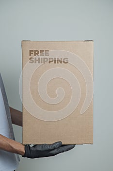 Detail of man with black gloves holding a cardboard box written free shipping to deliver products on gray background.