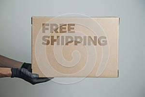 Detail of man with black gloves holding a cardboard box written free shipping to deliver products on gray background.