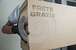 Detail of man with black gloves holding a cardboard box written free shipping to deliver products on gray background.
