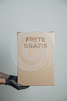 Detail of man with black gloves holding a cardboard box written free shipping to deliver products on gray background.