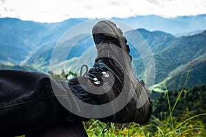 Detail of a man's trekking shoe and leg at rest in front of the green mountain landscape and the blue sky in the Himalayas.