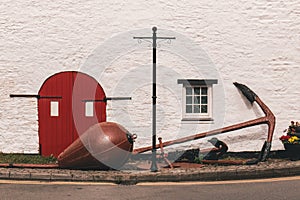 Detail of an old barn red door with black metal hinges against a white wall and a huge anchor outside