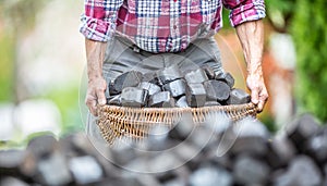 Detail of old man's arms picking up a basket full of coal briquettes with a blurred pile of briquettes in the front