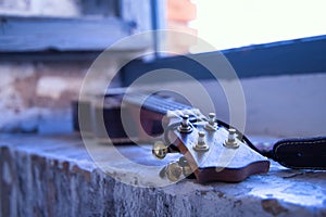 Detail of the pegbox, fret and strings of a brown ukulele with strap leaning against a brick window frame. Concept instruments,