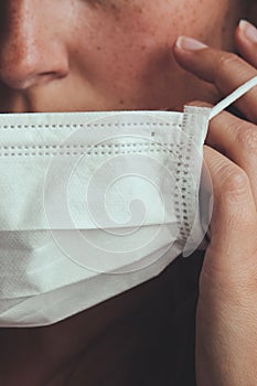 Detail photo of unrecognizable white woman putting on a white medical face mask to prevent the flu. Blurred background, focus on