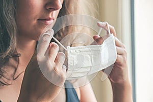 Detail photo of unrecognizable white woman putting on a white medical face mask to prevent the infection. Blurred background,