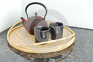 Detail of a red Chinese teapot and two black cups resting on a wicker tray, in a bright kitchen